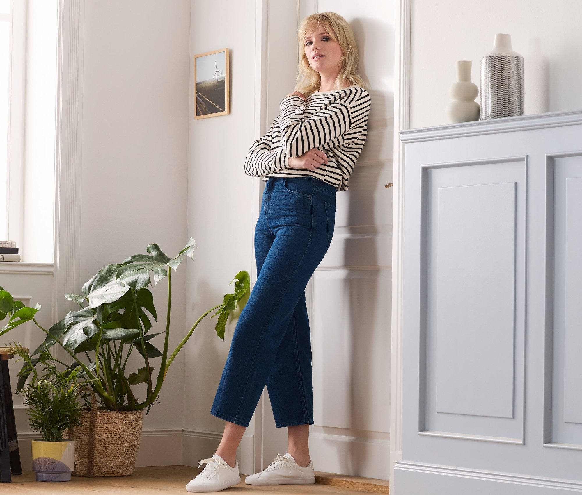 Eine Frau mit verschränkten Armen lehnt an einer Tür und trägt Jeans mit weitem Bein, ein gestreiftes Oberteil und Turnschuhe.