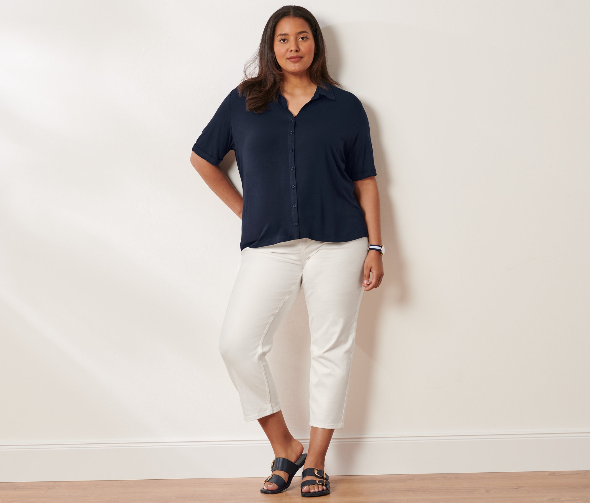 Frau posiert in Blusenshirt mit Knopfleiste, Slimfit-Jeans und Sandalen mit Schnalle. Sie trägt eine Quarz-Armbanduhr aus Edelstahl.