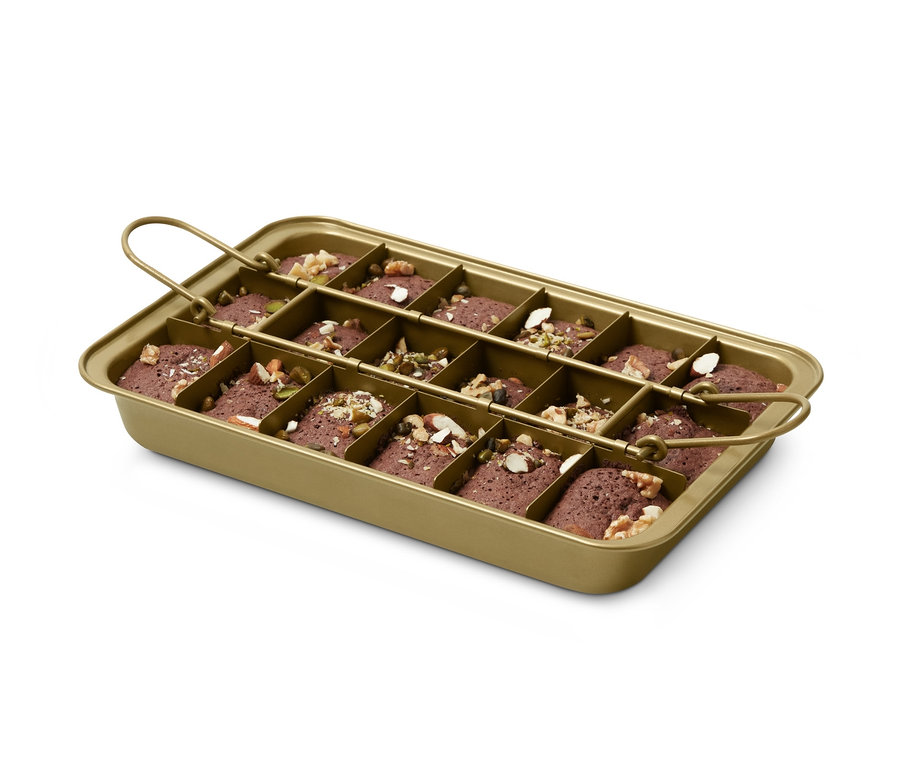 Eine goldene Brownie-Backform mit Griffen, gefüllt mit Teig und mit Nüssen und Samen bestreut.