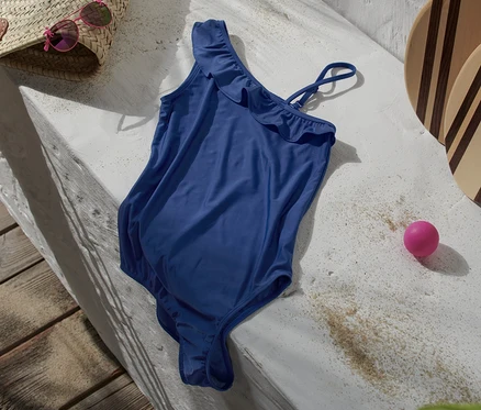 Blauer Badeanzug mit Rüschen liegt auf einer Betonfläche neben einer Strandtasche, einer Sonnenbrille, Strandpaddeln und einem rosa Ball.