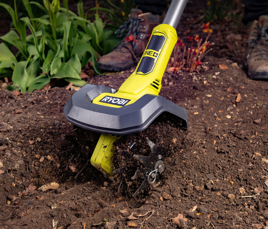 Gelbe RYOBI 18 V Akku-Bodenhacke »RY18CVA-0« lockert Erde in einem Gartenbeet, im Hintergrund sind die braunen Schuhe einer Person zu sehen.