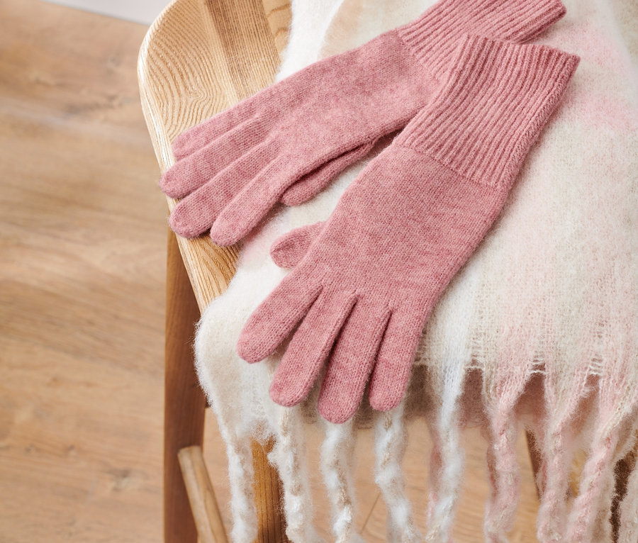 Rosafarbene Strickhandschuhe mit Wolle liegen auf einem Holzstuhl mit einem weißen Schal.