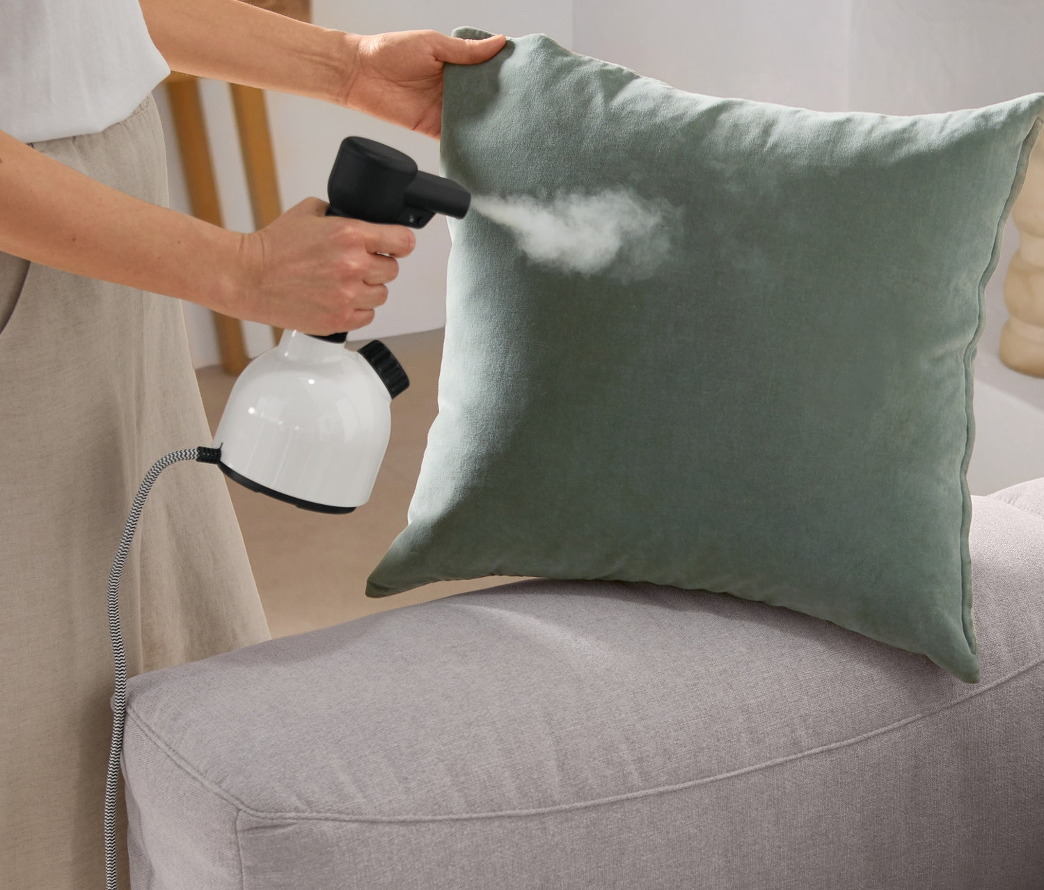 Frau dämpft ein grünes Kissen auf einem grauen Sofa mit einem Laurastar Hygiene-Steamer »IGGI Pure White«.