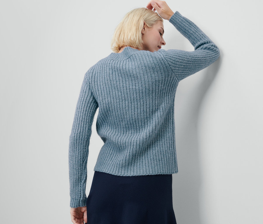 Frau mit blonden Haaren hat eine Hand am Kopf und trägt einen hellblauen Grobstrick-Cardigan.