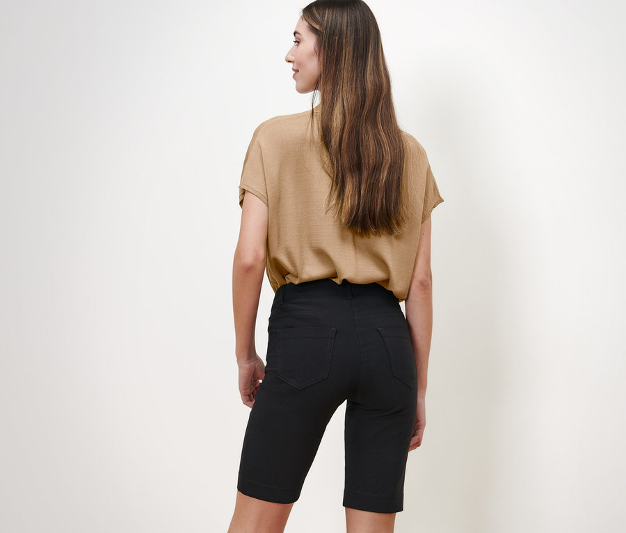 Eine Frau steht mit dem Rücken zur Kamera und trägt eine beige Bluse und schwarze Bengalin-Bermudashorts.