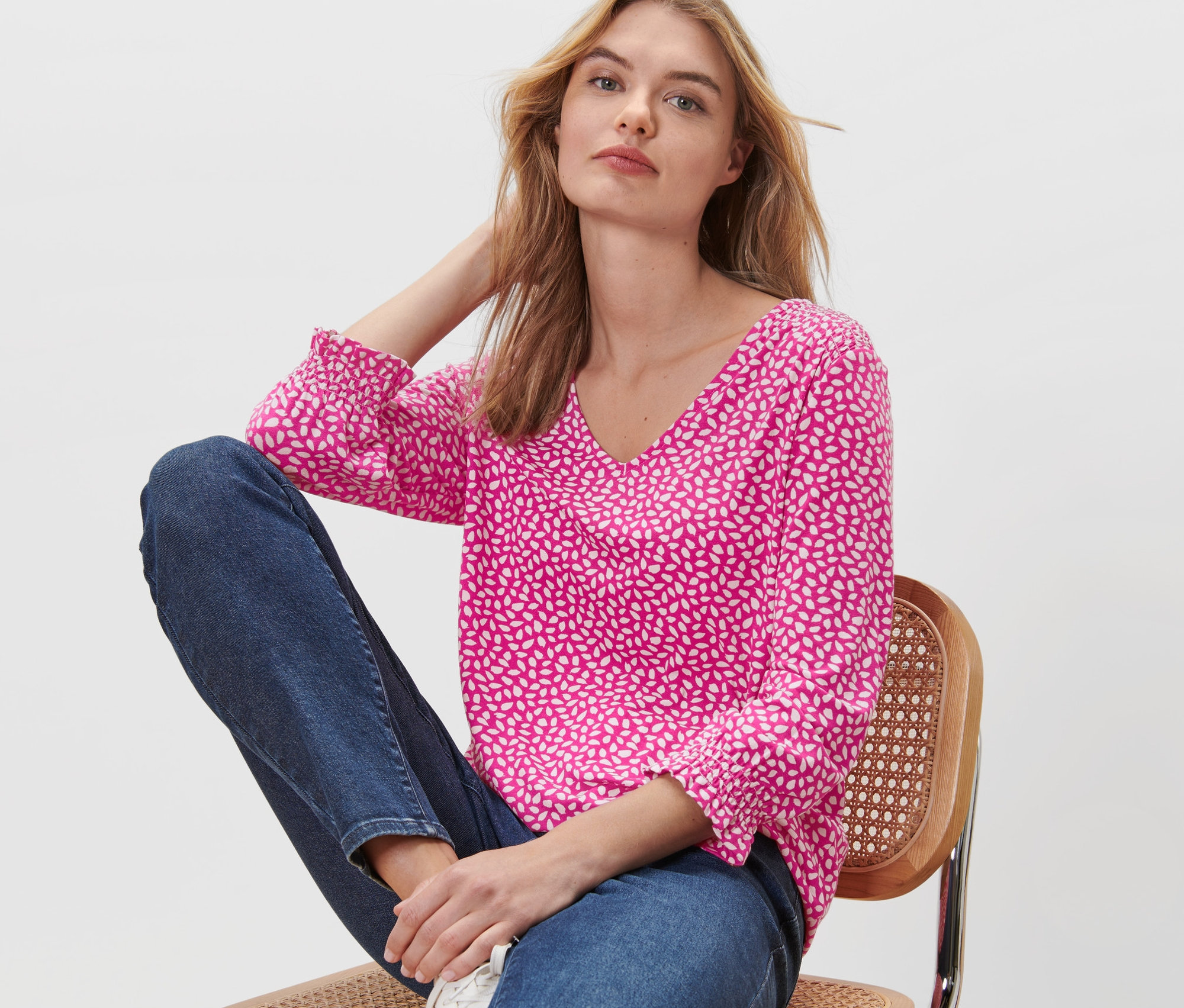 Frau sitzt auf einem Stuhl und trägt ein bedrucktes Blusenshirt in Pink.