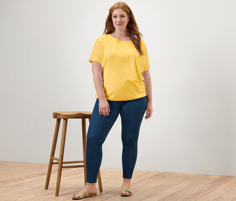 Frau posiert neben einem Hocker und trägt ein gelbes Blusenshirt mit Stickerei und Jeggings.