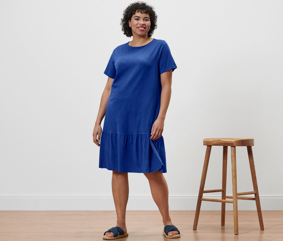Eine Frau steht und trägt ein kobaltblaues Jerseykleid mit Volant und Komfortsandalen.