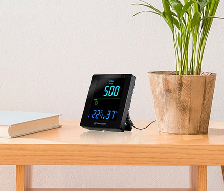 Auf einem Tisch stehen ein BRESSER CO2-Luftqualitätsmonitor Smile mit CO2-Ampel, ein Buch und eine Topfpflanze.
