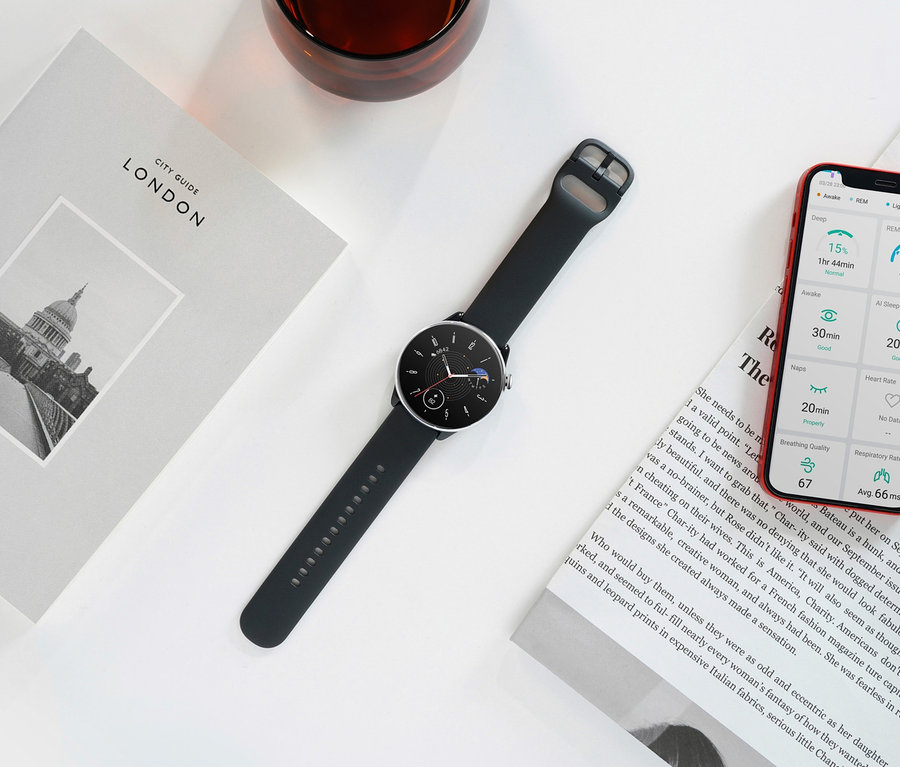Eine schwarze Amazfit GTR Mini Smartwatch liegt auf einer weißen Oberfläche neben einem Buch über London, einem Glas und einem Mobiltelefon.