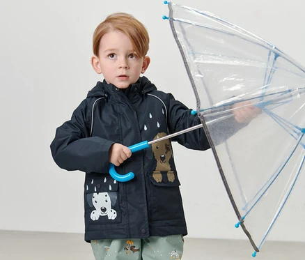 Junge hält durchsichtigen Regenschirm und trägt Kinder-Thermo-Regenjacke, Hund.