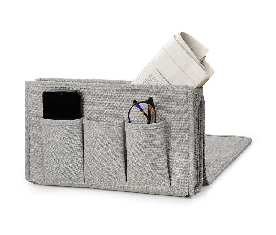 Ein grauer Bett-und-Couchorganizer mit Taschen, in denen sich ein Telefon, eine Brille und eine Zeitung befinden.