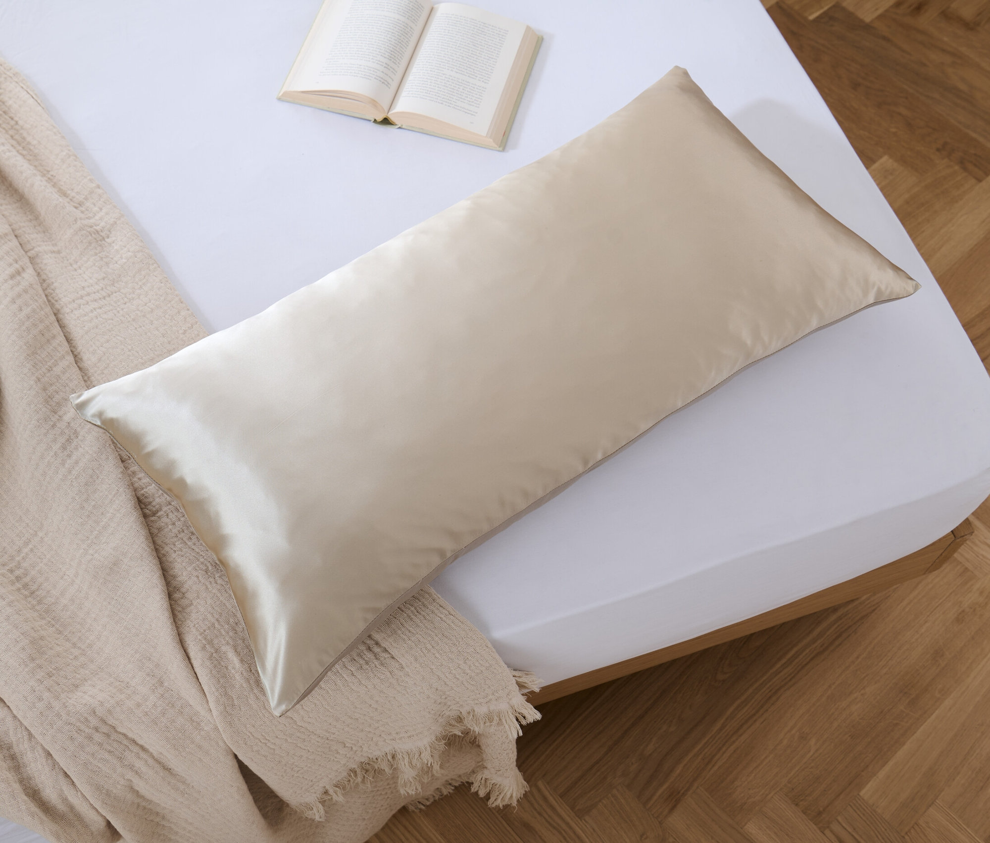 Ein beiger Seiden-Baumwoll-Kopfkissenbezug, ca. 100 x 65 cm, liegt auf einem Bett neben einem offenen Buch und einer Tagesdecke.