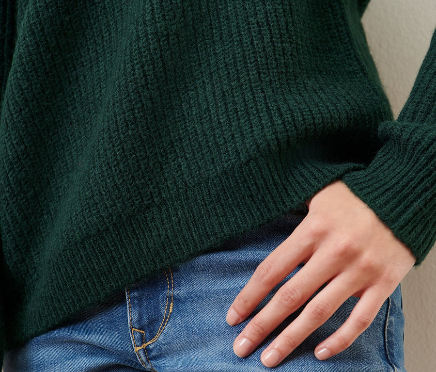 Detail einer Frau, die einen dunkelgrünen Strickpullover mit Stehkragen und blaue Jeans trägt.