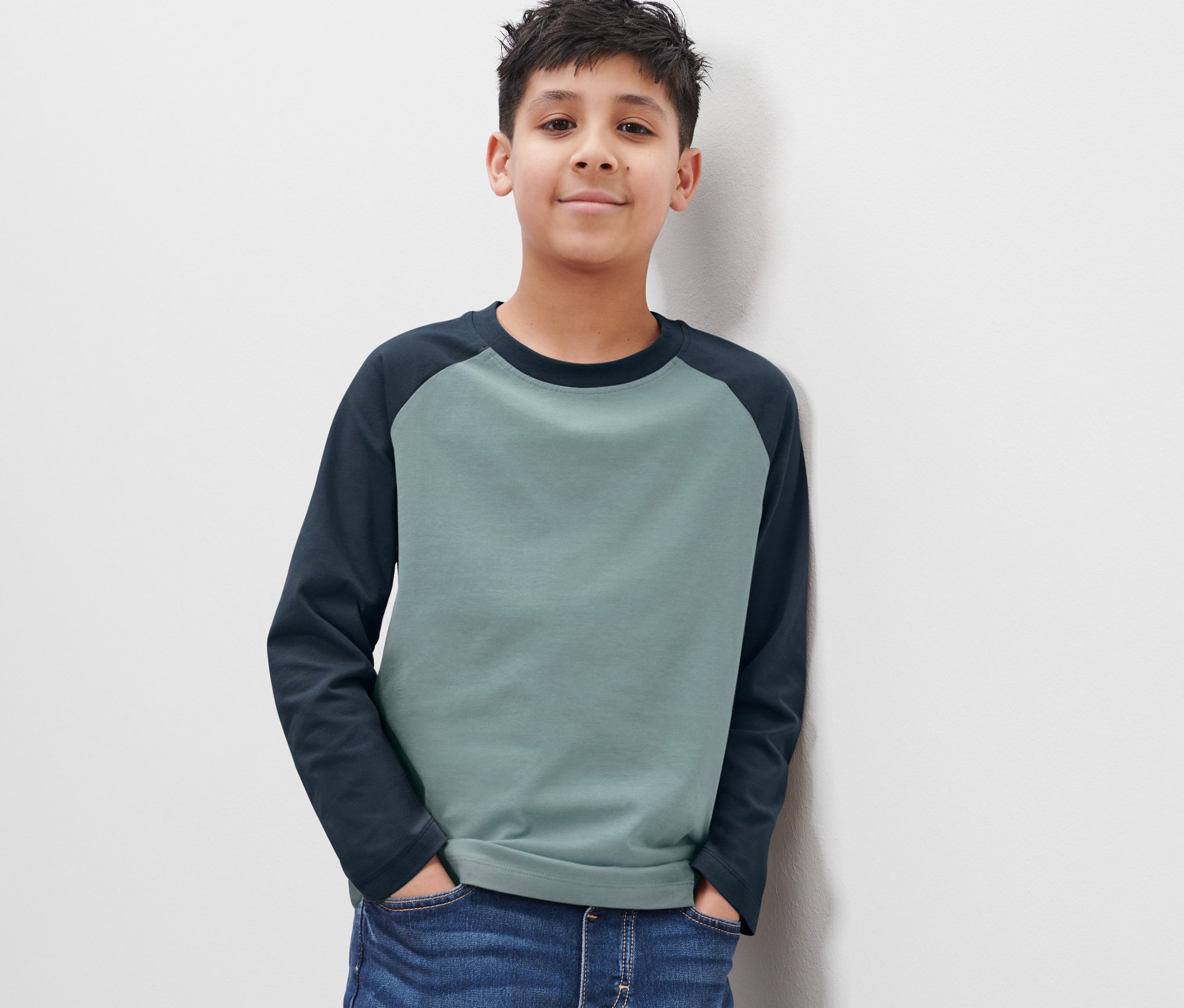 Ein Junge in blauen Jeans und einem zweifarbigen Langarmshirt steht mit dem Rücken zu einer weißen Wand.
