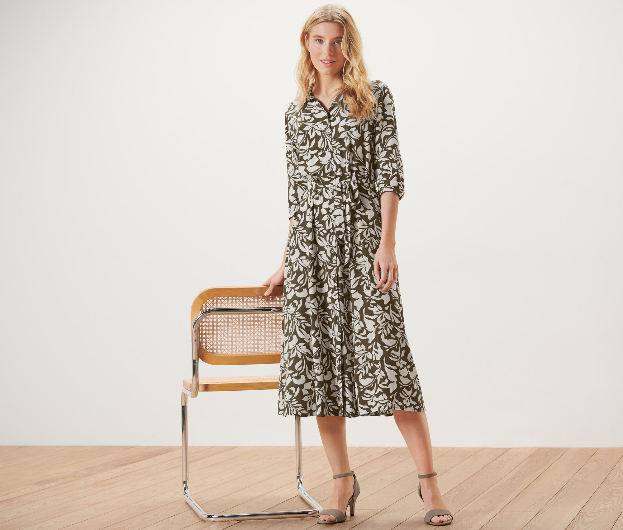 Blonde Frau posiert neben einem Stuhl und trägt ein bedrucktes Hemdblusenkleid.