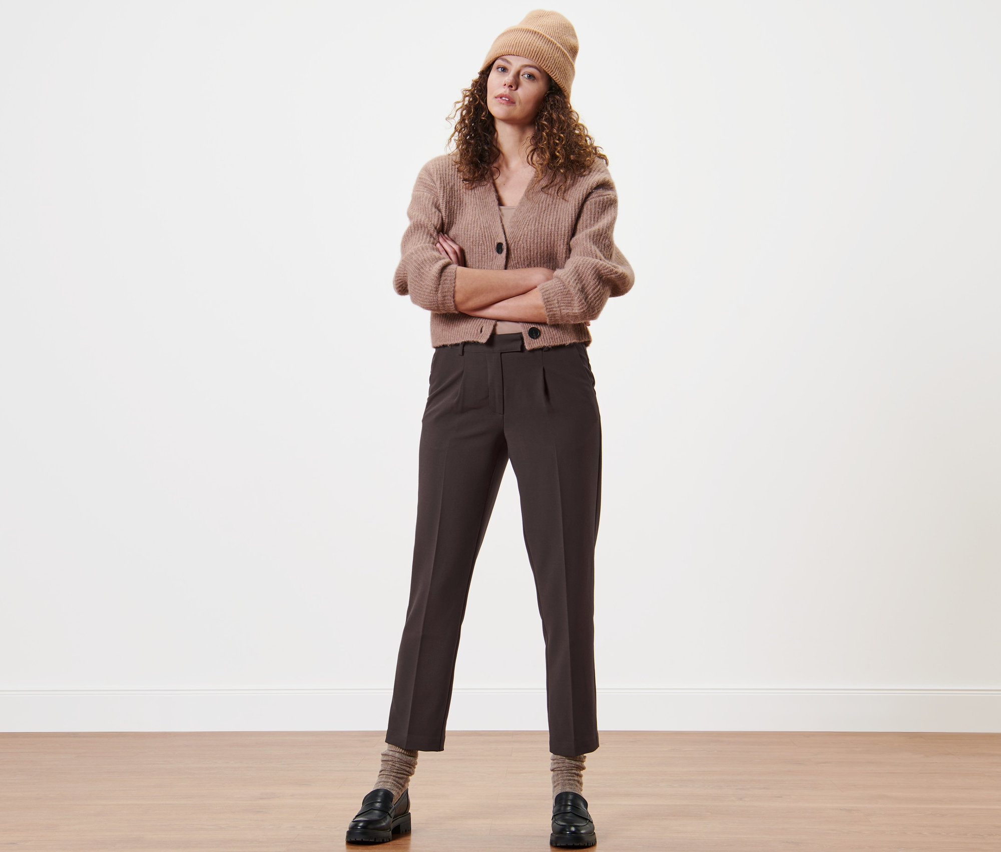 Frau mit verschränkten Armen steht und trägt eine Webhose mit Bundfalten, eine Strickjacke und eine Mütze.