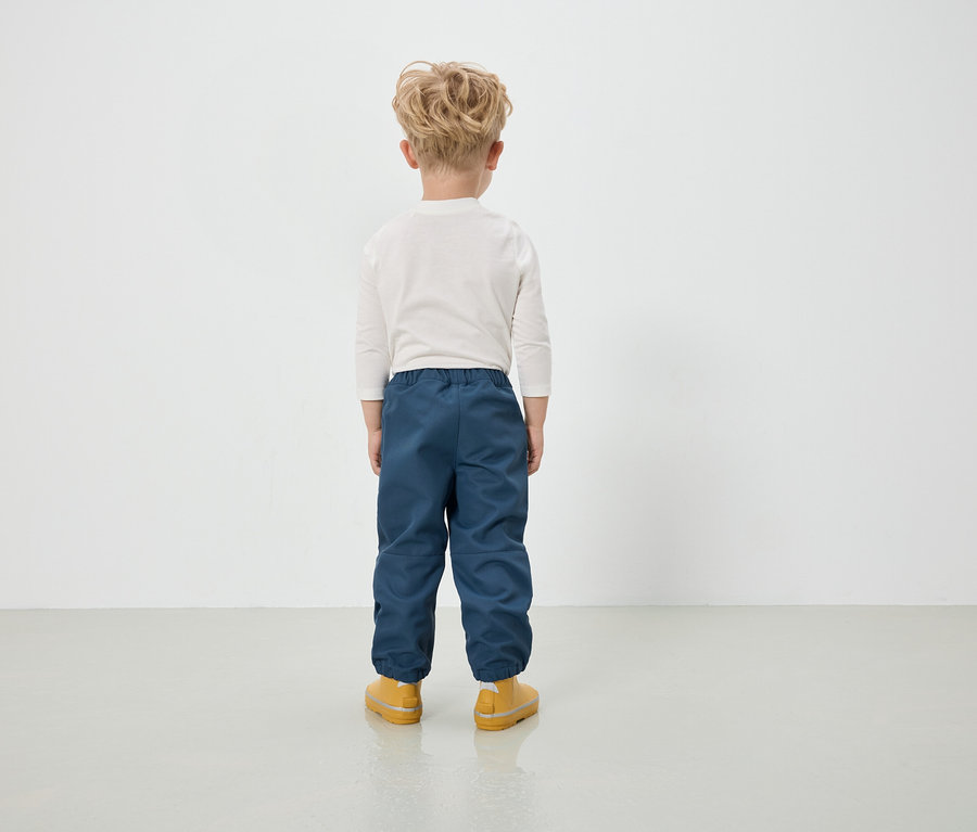 Junge in blauen Hosen und gelben Stiefeln steht mit dem Rücken zur Kamera.