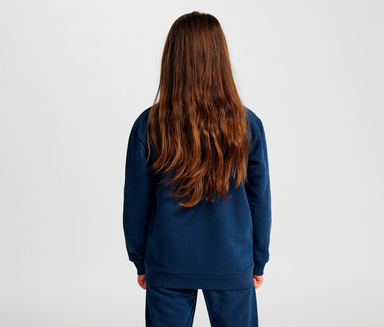 Mädchen mit braunen Haaren steht mit dem Rücken zur Kamera und trägt ein blaues HUMMEL HMLJR Base-Sweatshirt.