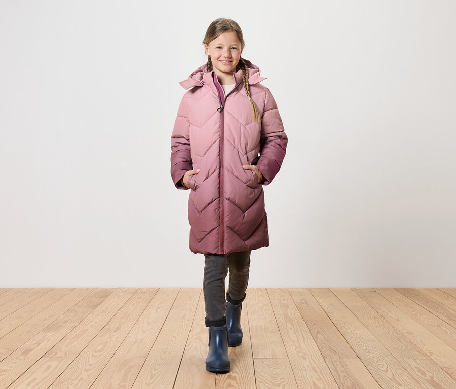 Ein Mädchen mit blonden Zöpfen geht über einen Holzboden und trägt einen langen, gesteppten Mantel mit Kapuze, der von Rosa zu Lila übergeht.