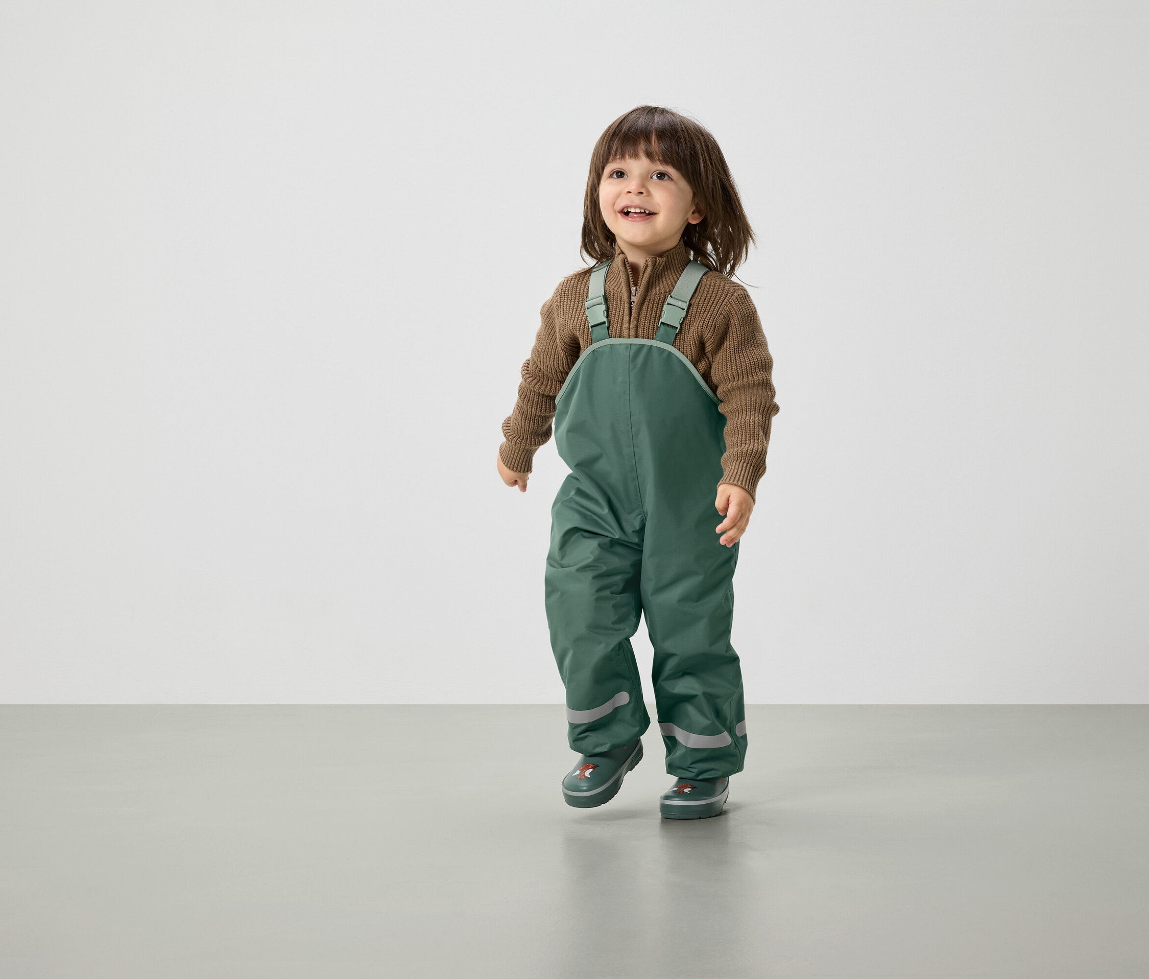 Lächelndes Kind mit braunen Haaren geht in grüner Latzhose und braunem Pullover.