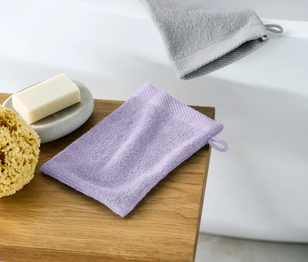 Auf einem Holzhocker liegen ein lila Waschhandschuh, daneben ein Naturschwamm und eine graue Seife auf einer Untertasse. Im Hintergrund sind der Rand einer Badewanne und ein grauer Waschhandschuh zu sehen.