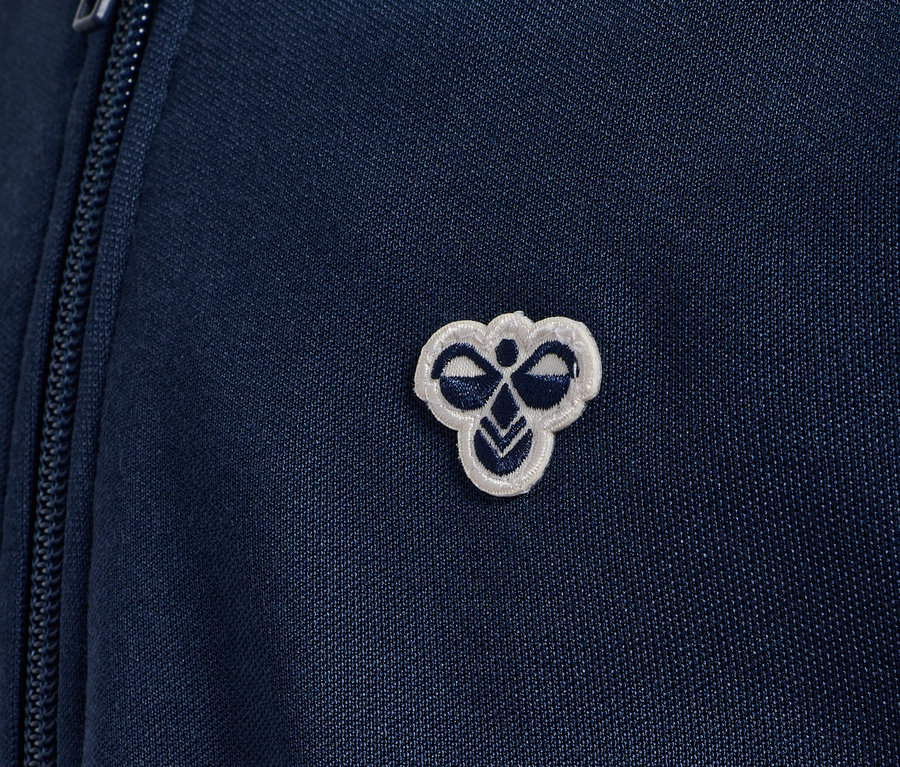 Detailansicht einer blauen Hummel HMLJR Archive Reg Poly Zip Jacket mit Logo.