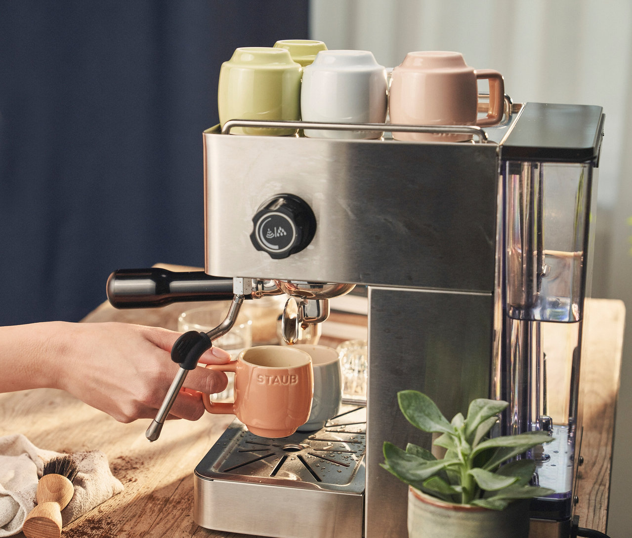 Eine Hand befüllt eine STAUB Espressotasse »Macaron« mit einer Espressomaschine. Auf der Maschine stehen weitere Tassen in Weiß, Grün und Rosa.