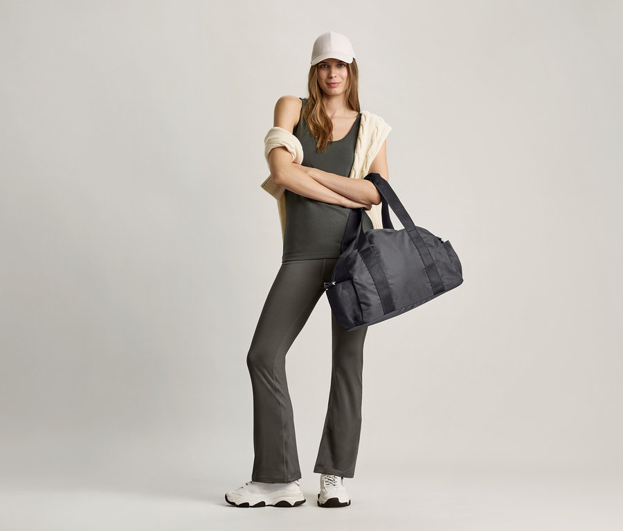 Frau steht mit gekreuzten Armen und hält eine schwarze Sporttasche. Sie trägt ein olivgrünes Top und eine graue Hose.