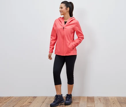 Eine lächelnde Frau posiert in einer korallenroten 2,5-Lagen-Outdoorjacke, einer schwarzen Dreiviertel-Leggings und Wanderschuhen.