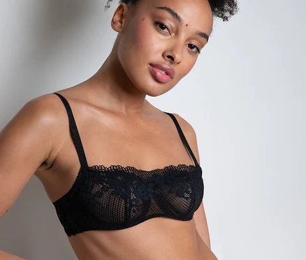 Eine Frau mit hochgesteckten Locken trägt einen schwarzen Passionata Bandeau-BH mit Spitze »Nina« und passende Höschen, eine Hand an der Hüfte.