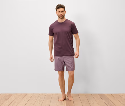 Mann steht barfuß auf einem Holzboden und trägt ein weinrotes T-Shirt und lila Shorts.