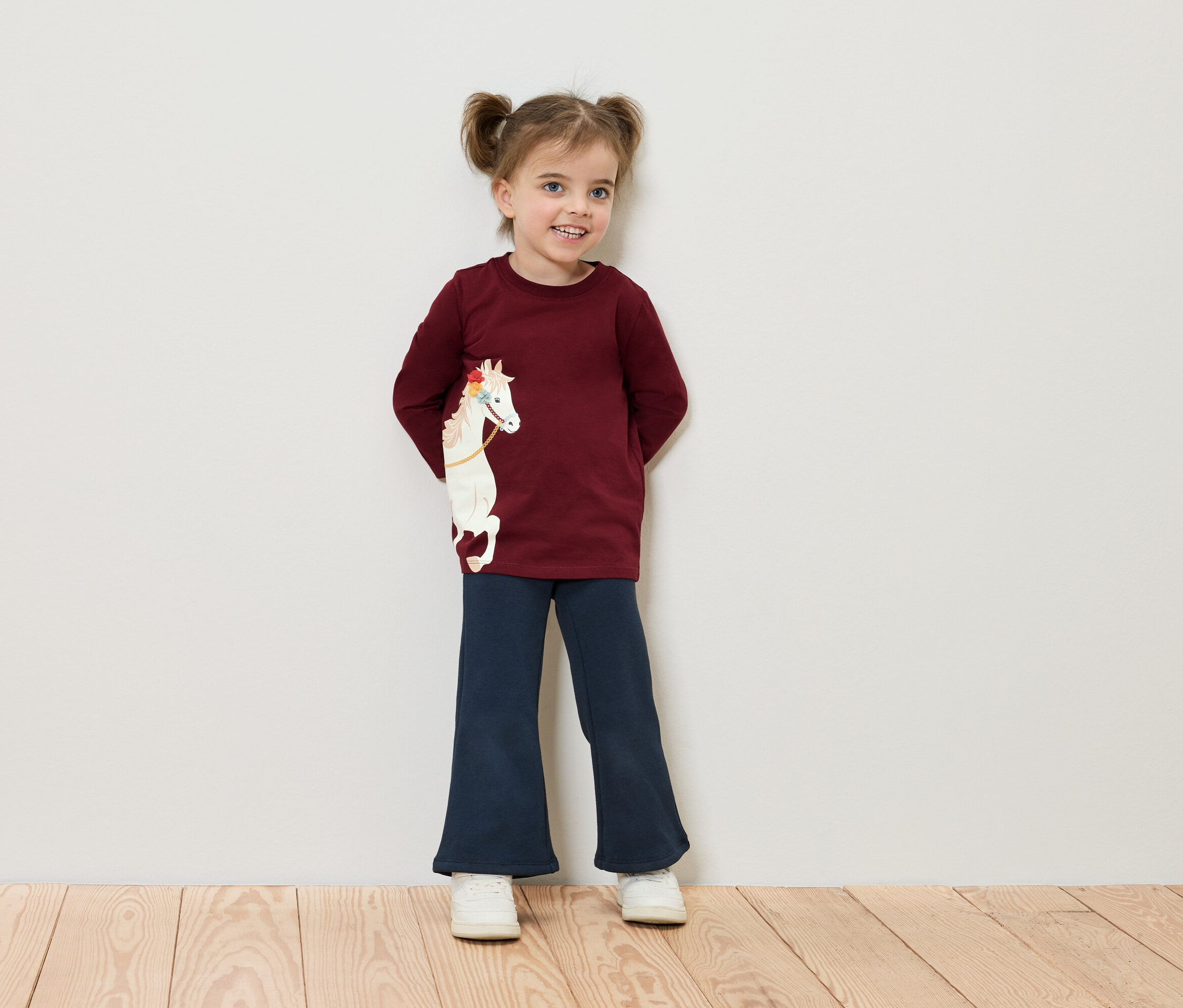 Lächelndes kleines Mädchen mit Zöpfen steht an einer Wand und trägt ein burgunderrotes Hemd mit einem Pferd und eine blaue Hose.