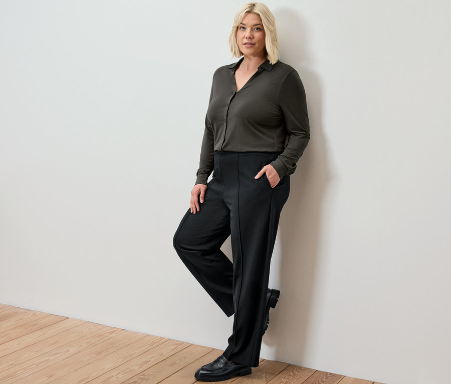 Blonde Frau, die sich an eine Wand lehnt, trägt eine schwarze Jerseyhose mit Biese und ein Blusenshirt mit Knopfleiste.