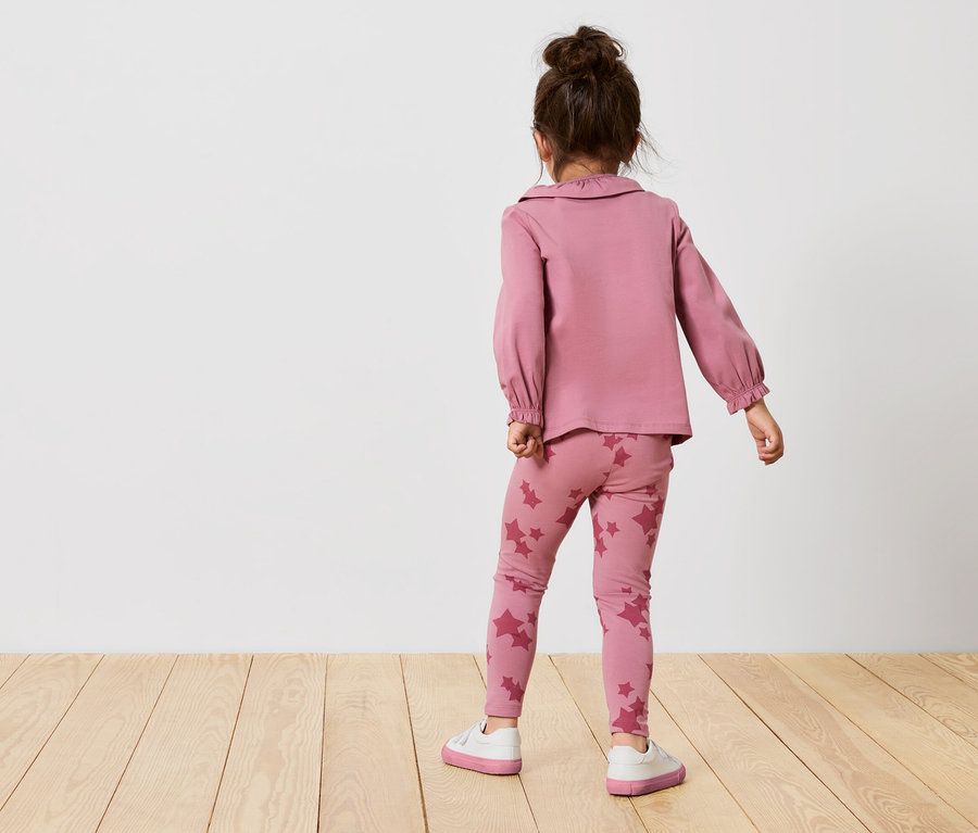 Mädchen in rosafarbenem Sternen-Ensemble steht mit dem Rücken. Sie trägt ein rosa langärmliges Oberteil und Leggings mit Sternen.