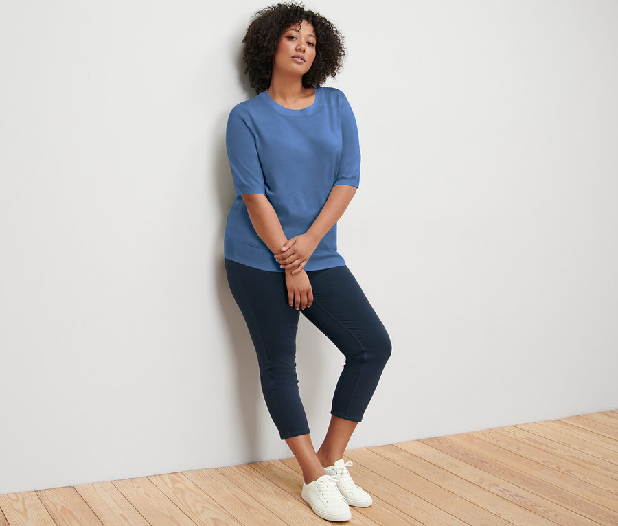 Eine Frau lehnt an einer Wand und trägt einen blauen Feinstrickpullover, dunkelblaue Hosen und weiße Turnschuhe.