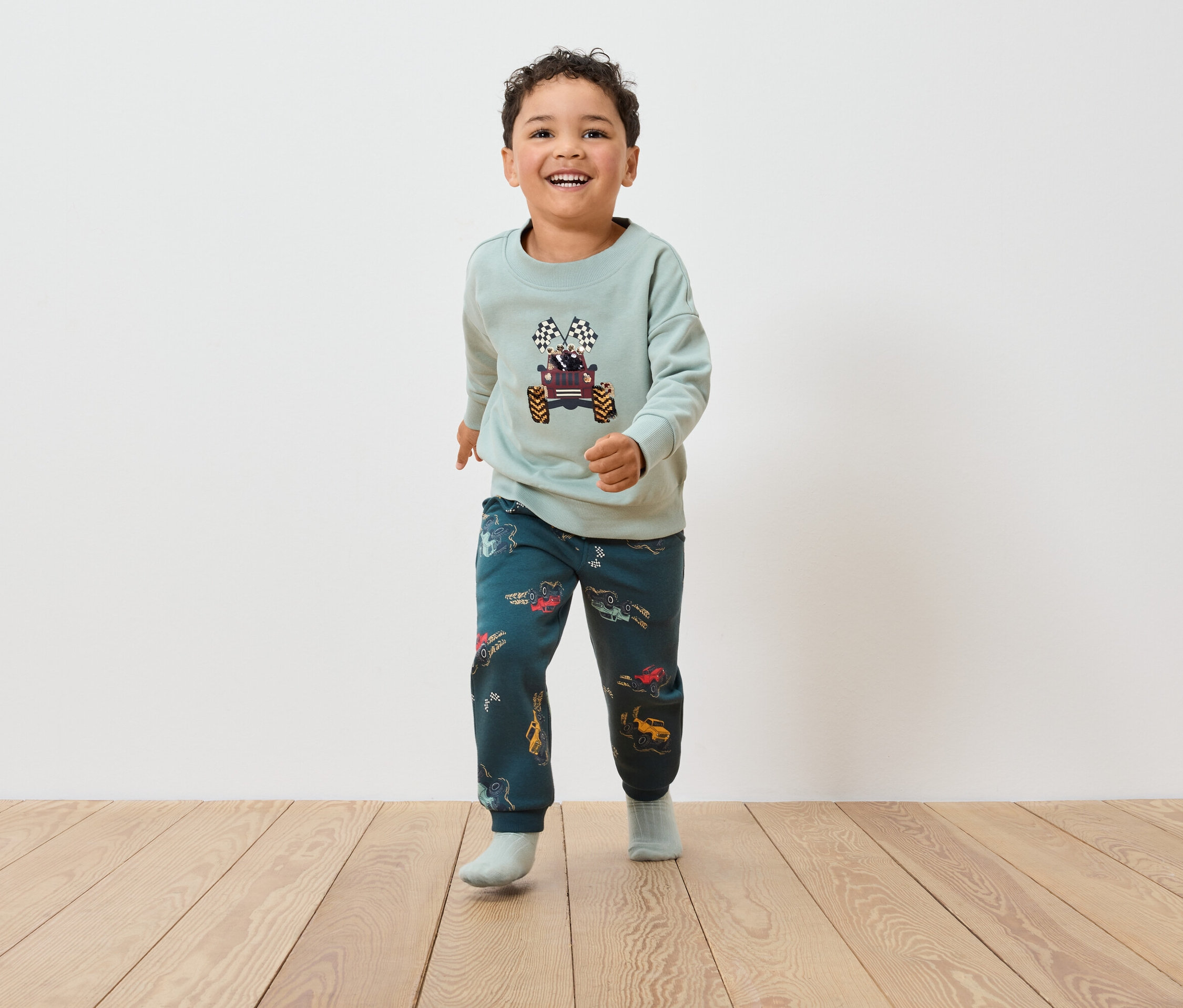 Ein Junge in einem Monster-Truck-Sweatshirt und Hosen mit Autoaufdruck geht auf einem Holzboden.