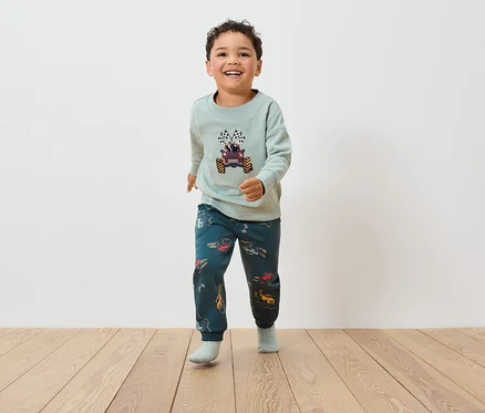 Ein Junge in einem Monster-Truck-Sweatshirt und Hosen mit Autoaufdruck geht auf einem Holzboden.