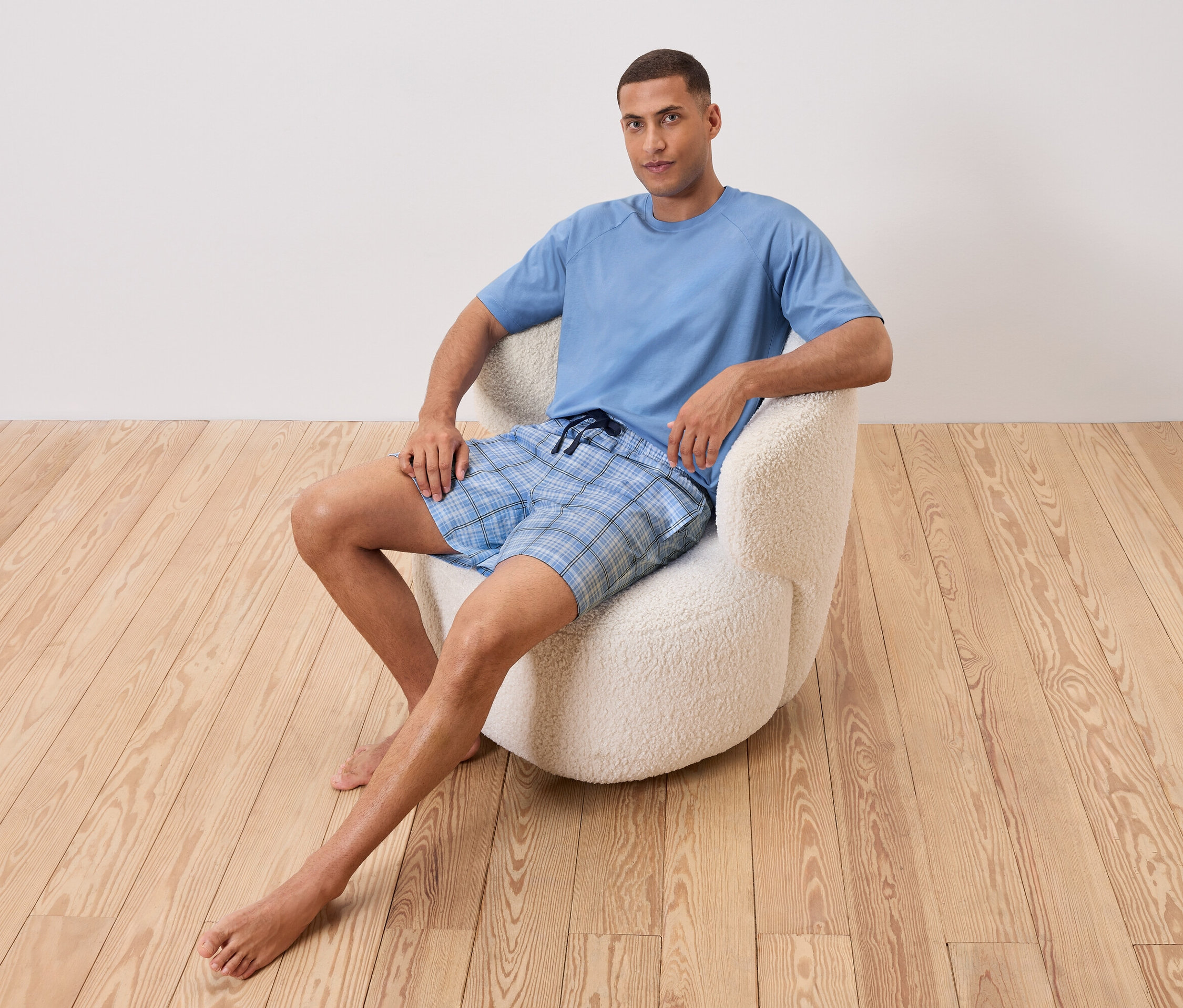 Ein Mann sitzt barfuß in einem weißen Sessel. Er trägt ein hellblaues T-Shirt und Shorts mit blauem Karomuster.