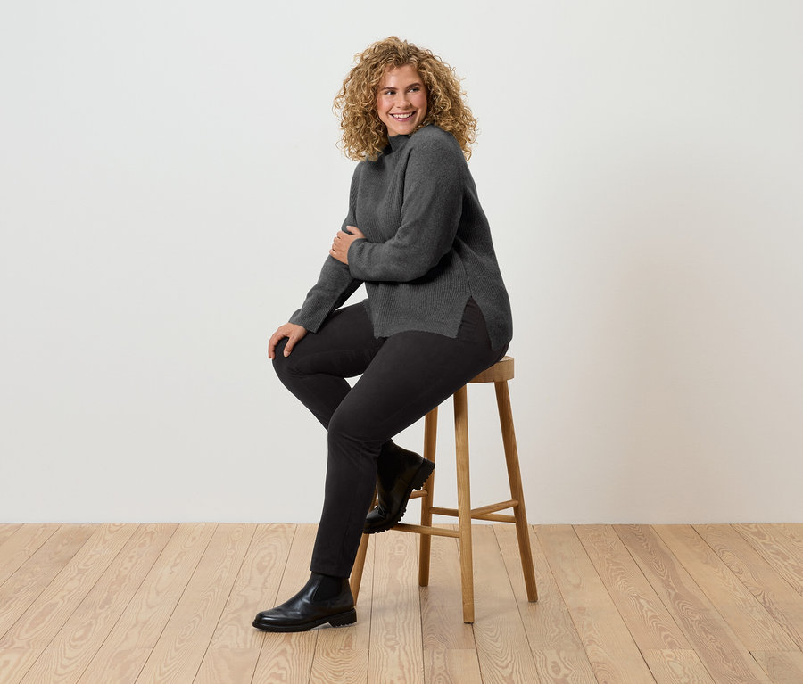 Eine Frau mit lockigem Haar sitzt auf einem Holzhocker. Sie trägt einen grauen Rollkragenpullover, schwarze Hosen und schwarze Stiefel.