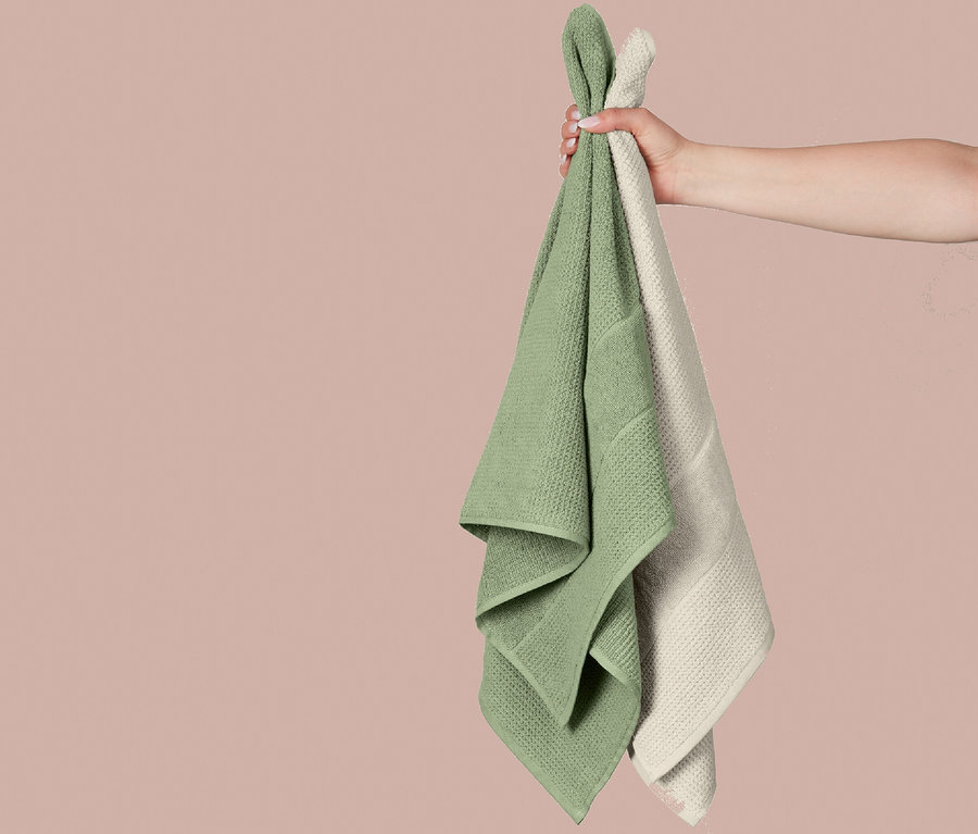 Eine Hand hält zwei SCHIESSER Duschtücher, ein grünes und ein hellbeiges, aus dem Turin Duschtuchset, vor rosa Hintergrund.