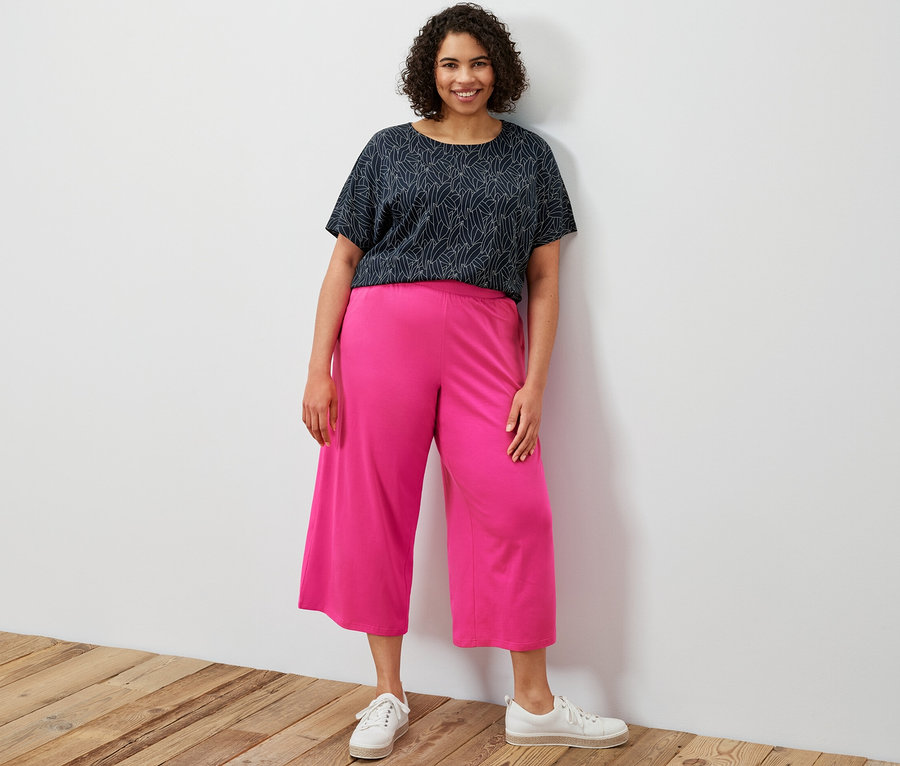 Eine Frau steht und trägt ein dunkelblaues, kurzärmeliges, überschnittenes Blusenshirt und eine pinke Jersey-Culotte.