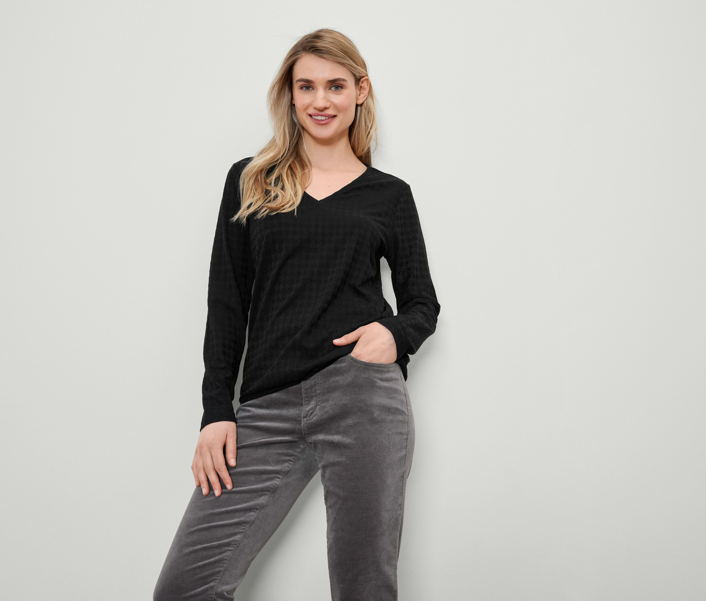 Blonde Frau in schwarzem Langarmpullover und grauer Cordhose steht und lächelt.