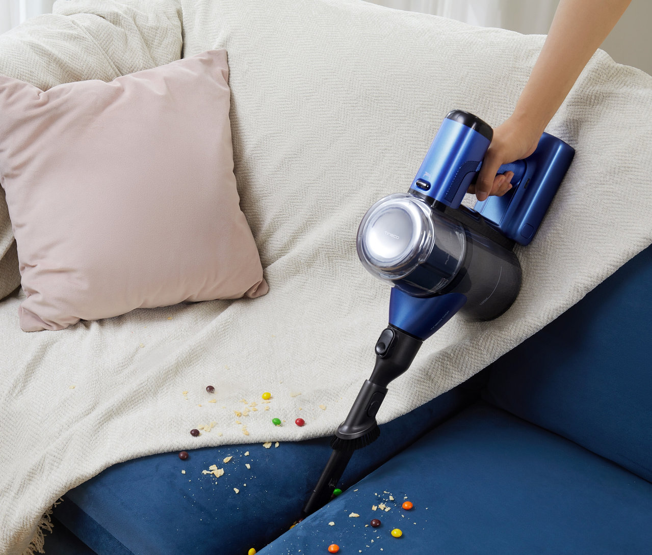 Eine Hand hält einen blauen Tineco Akku-Staubsauger »Pure One S50 Pro« über ein blaues Sofa mit Krümeln und Süßigkeiten.