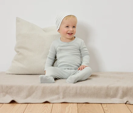 Ein lächelndes Kleinkind mit geringelter Mütze sitzt auf einer beige Decke vor einem Kissen.