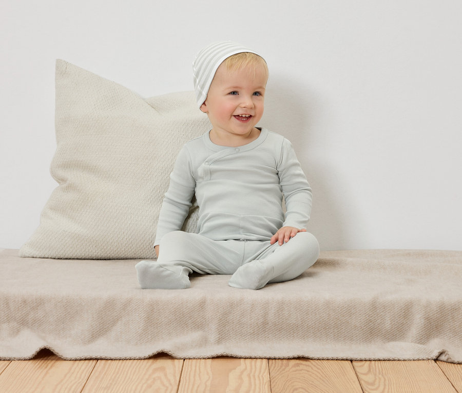 Ein lächelndes Kleinkind mit geringelter Mütze sitzt auf einer beige Decke vor einem Kissen.