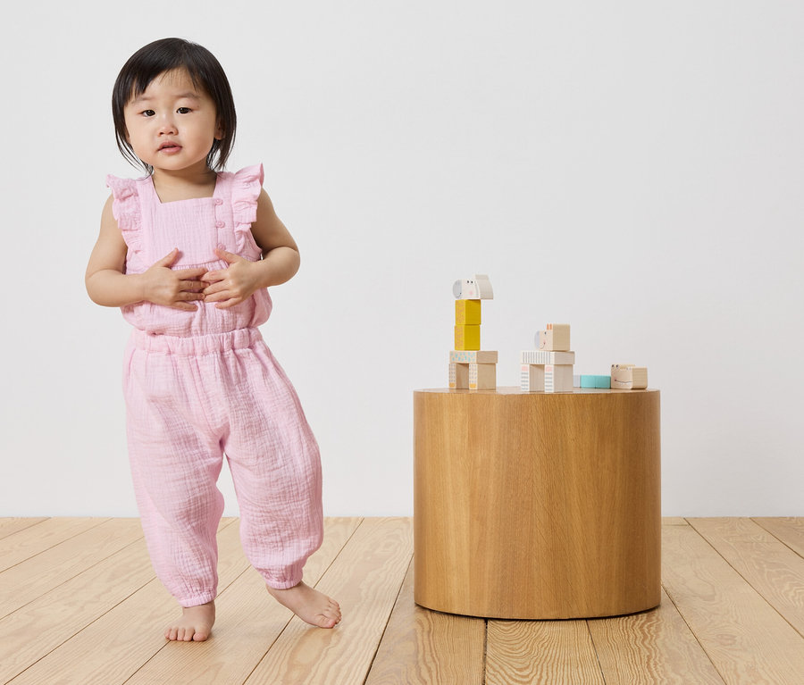 Kleines Mädchen in rosa Musselin-Jumpsuit steht neben einem runden Holztisch mit Bauklötzen.