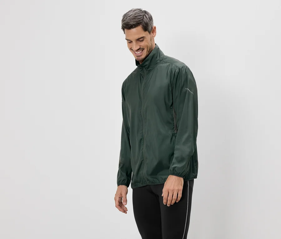 Mann in grüner Jacke und schwarzen Leggings steht vor einem weißen Hintergrund und lächelt.