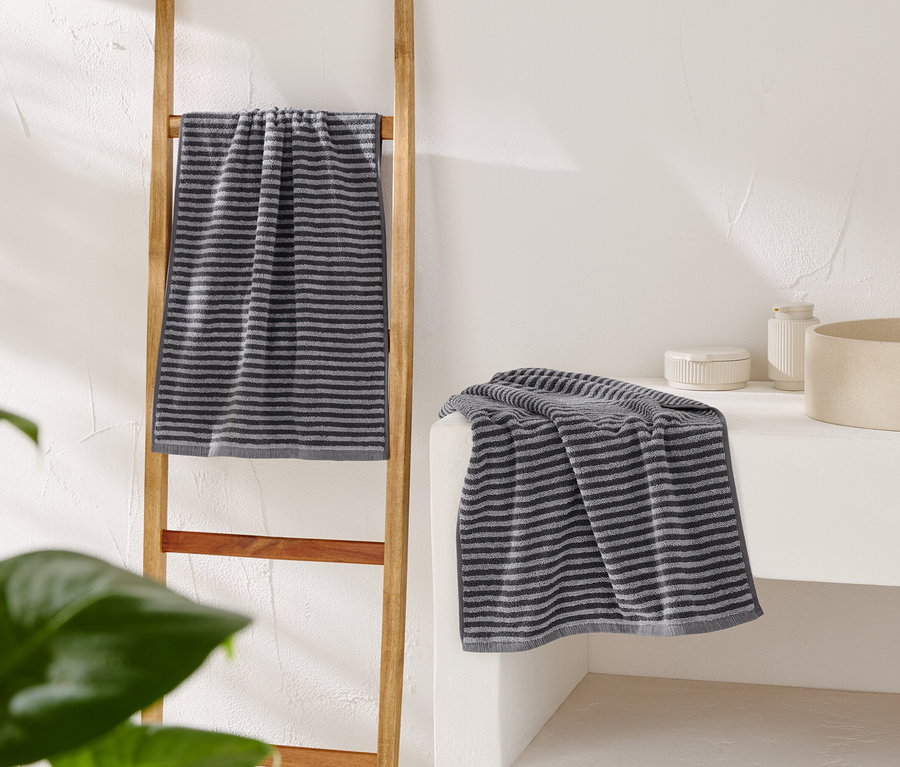 Ein gestreiftes graues Handtuch hängt an einer Holzleiter, ein weiteres liegt auf einem weißen Waschtisch neben einem beige Waschbecken.