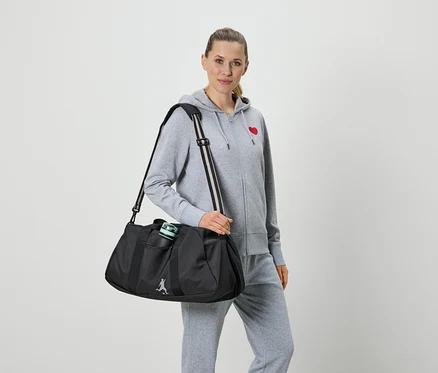 Frau mit grauer Trainingsjacke mit rotem Herz und grauer Trainingshose hält eine schwarze Sporttasche mit Trinkflasche.